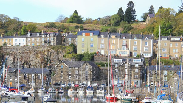 A harbor with multiple boats docked, surrounded by stone buildings with multiple stories and a hillside in the background. - Captain's Rest (Porthmadog)