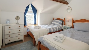 A bedroom with a sloped ceiling, a bed with a striped bedspread, a bedside table with a lamp, a dresser with drawers, and a window with curtains. - Captain's Rest (Porthmadog)