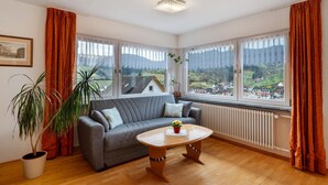Ein modernes Wohnzimmer mit grauem Sofa, einem Holztisch, einem Blumentopf und großen Fenstern mit Blick auf eine Stadt und Berge.