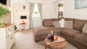 A living room with a brown sofa, a small wooden table with a bottle of wine, glasses, and pastries. There is a white cabinet with drawers, a lamp, and a vase with flowers on a side table. A window with curtains and a painting on the wall is visible. - Loft Cottage (Bedale)