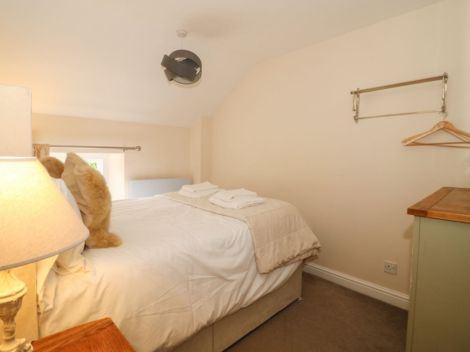 A bedroom with a bed, a bedside table, a lamp, a hanging light fixture, a coat rack, and a window with curtains.
