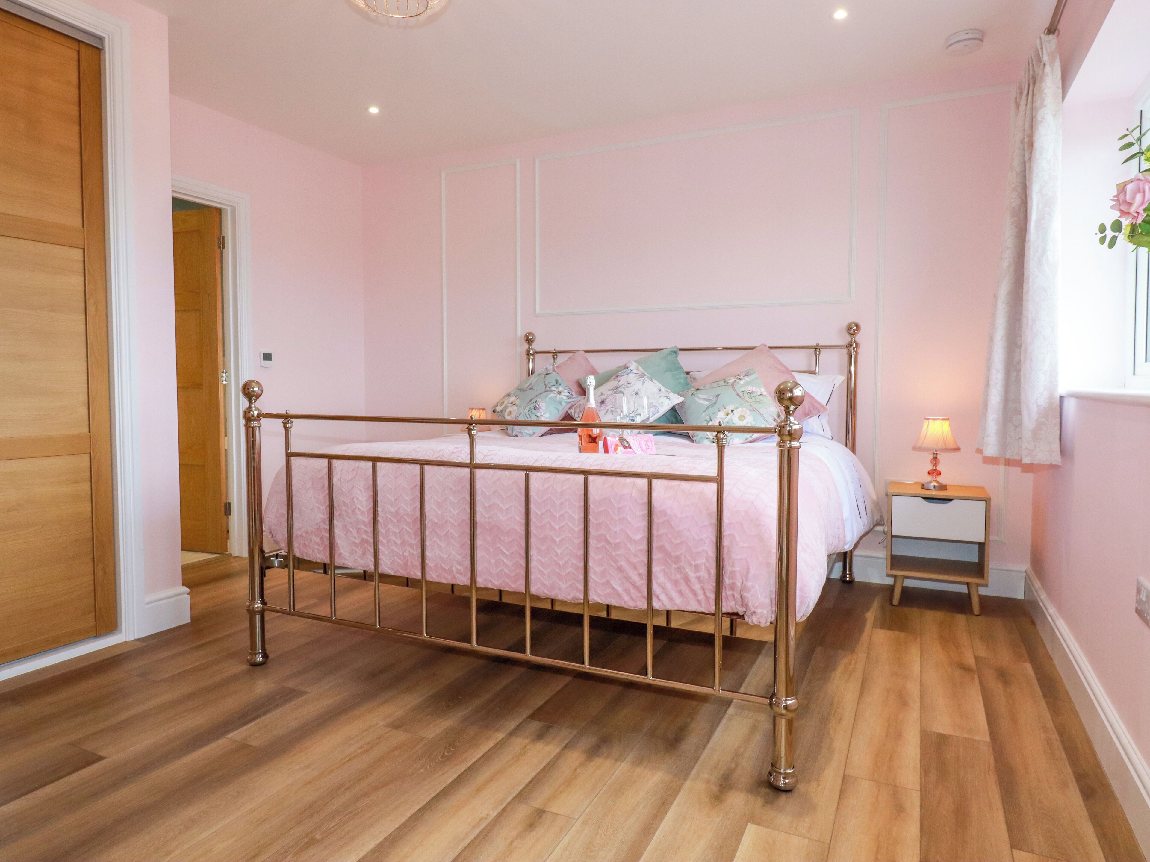 A bedroom with a wooden floor, a bed with a metal frame, a bedside table with a lamp, and a door.
