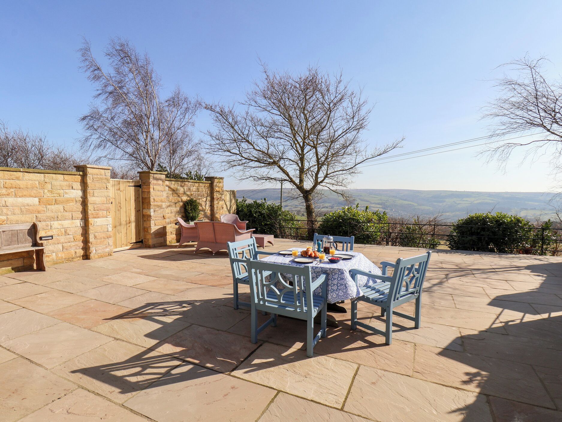 A patio with stone flooring, a table set with plates, and chairs. There is a wooden bench and a stone wall in the background.