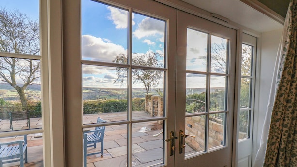 A room with a large window offering a view of a patio with a bench and a stone wall. - Roding Cottage (Whitby)