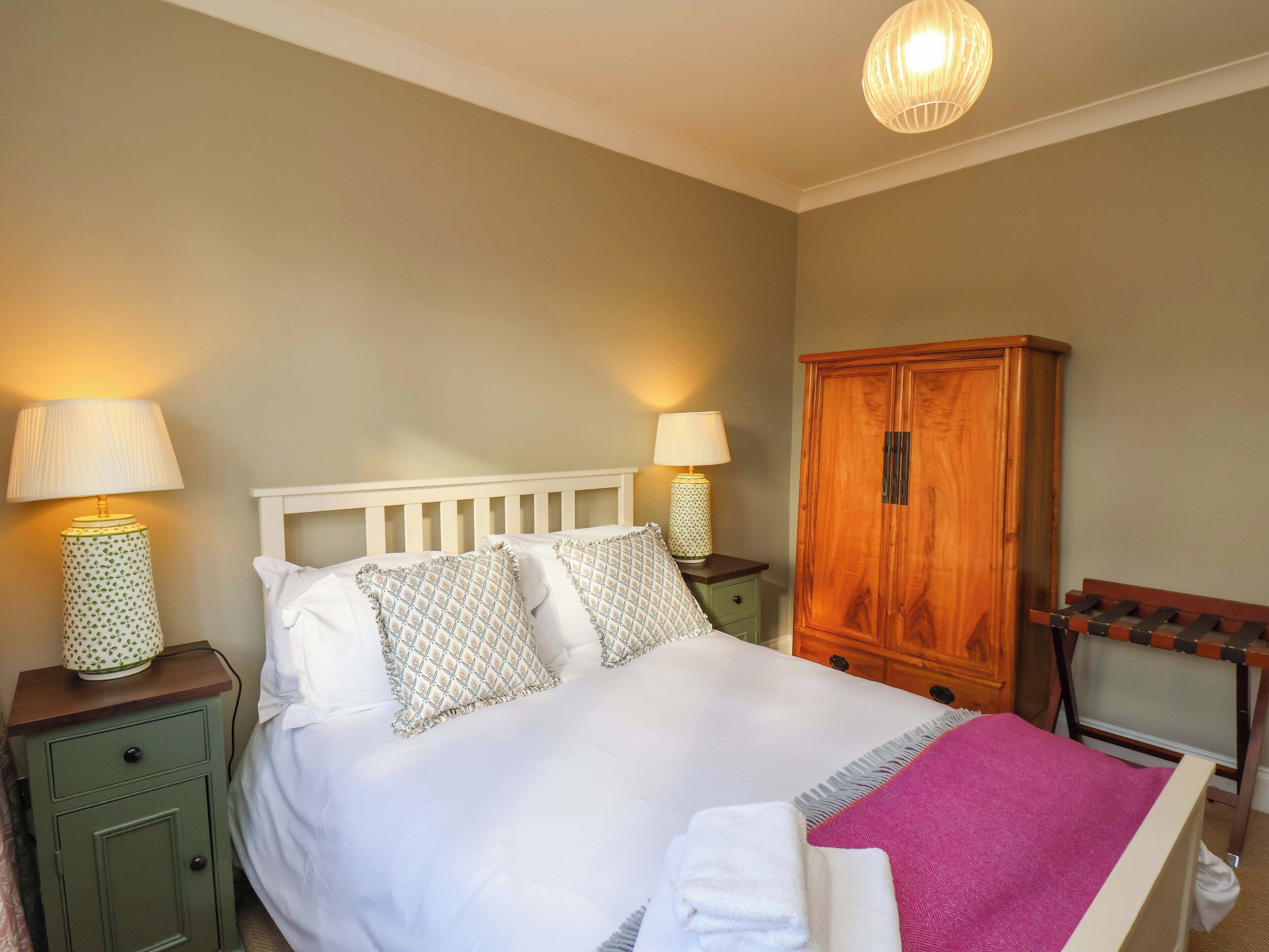 A neatly made bed with white linens and patterned pillows, flanked by two bedside tables with lamps, a wooden wardrobe, and a chair with a backrest.