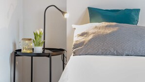 Una cama bien hecha con un cojín estampado y uno turquesa, una lámpara de noche, una planta pequeña y un tarro con etiqueta.