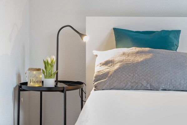 A neatly made bed with a patterned pillow and a teal pillow, a bedside lamp, a small plant, and a jar with a label.