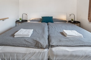 A double bed with grey bedding and white pillows, two bedside tables with lamps, and a small plant on each.