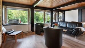 Un interior moderno con techo de madera, ventanas amplias, un sofá de cuero negro, una silla de madera y un reposapiés negro.