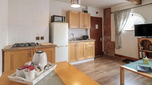 A compact kitchen with a gas stove, microwave, and refrigerator. There's a wooden table with a tray of items, a window with curtains, and a bookshelf with books. - Stables (Derby)