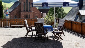 Un patio con sillas de mimbre, una mesa y un paraguas a rayas. Al fondo, se observa un edificio de ladrillo con una torre y una aguja de iglesia.