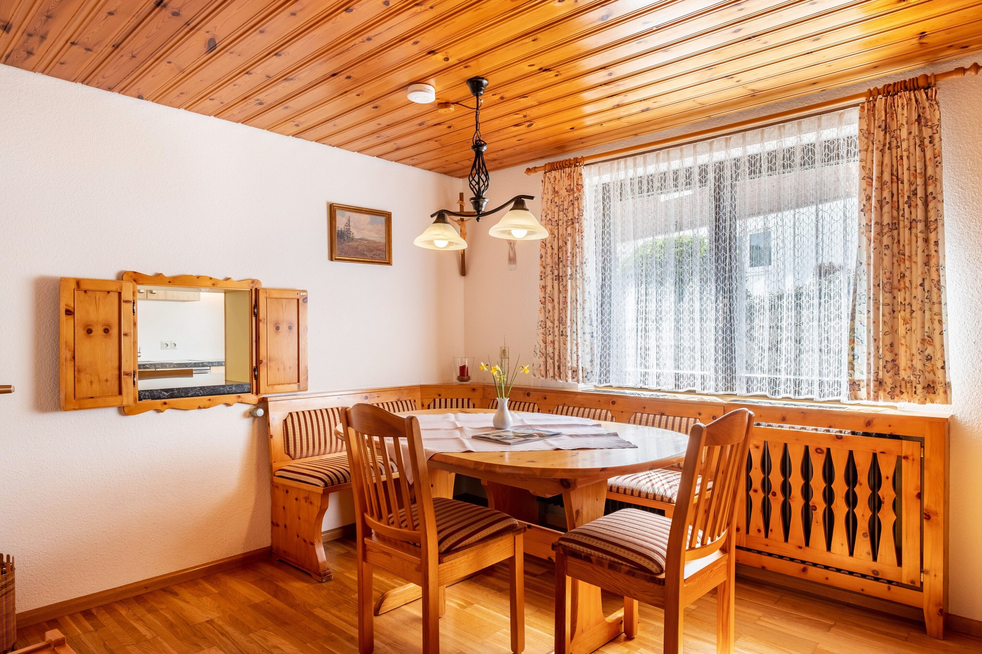A wooden dining room with a round table, chairs, a chandelier, and a window with curtains.