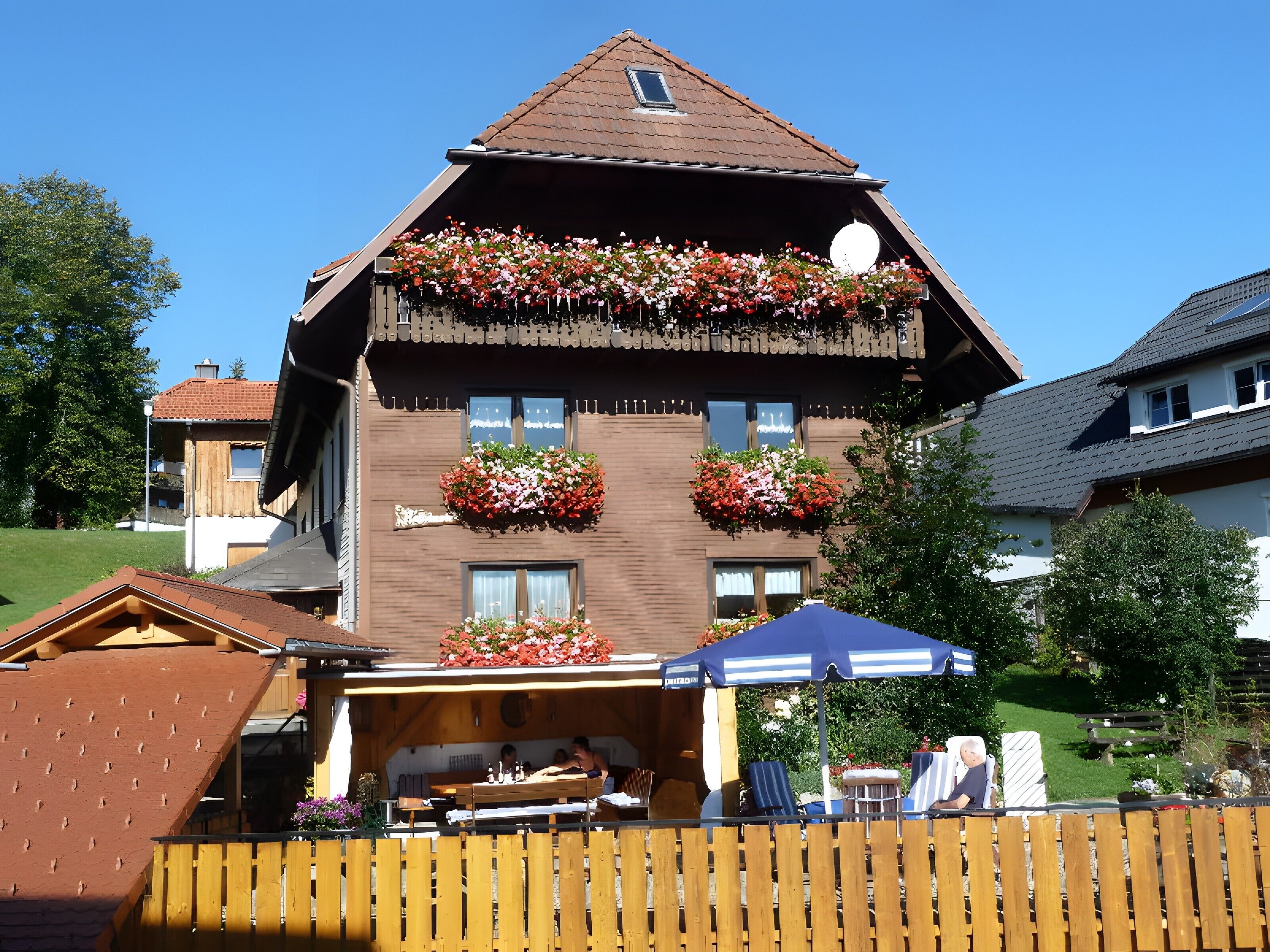 A two-story building with a balcony adorned with flowers, a wooden deck with tables and chairs, and a clear blue sky.