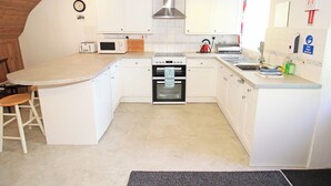 A compact kitchen with white cabinets, a built-in oven, a microwave, and a kettle. There is a sink with a hand sanitizer dispenser and a clock on the wall. - Y Beudy (Llanbedr)