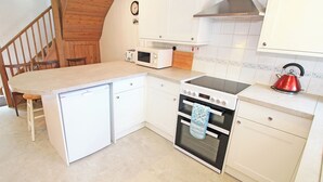A compact kitchen with white cabinets, a built-in oven, a microwave, and a red kettle. - Y Beudy (Llanbedr)