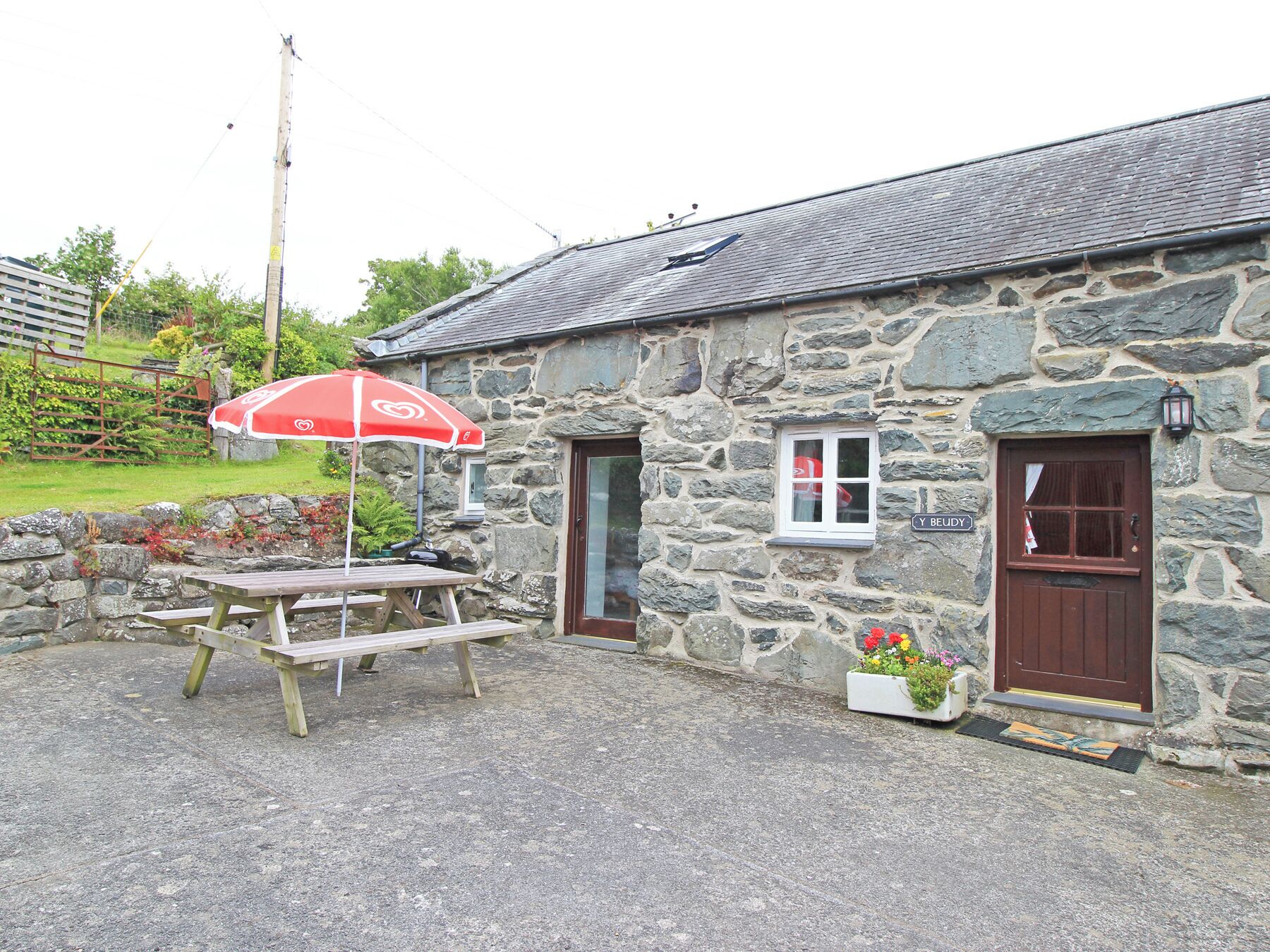 A stone building with a wooden door and window, a picnic table, and a red umbrella.