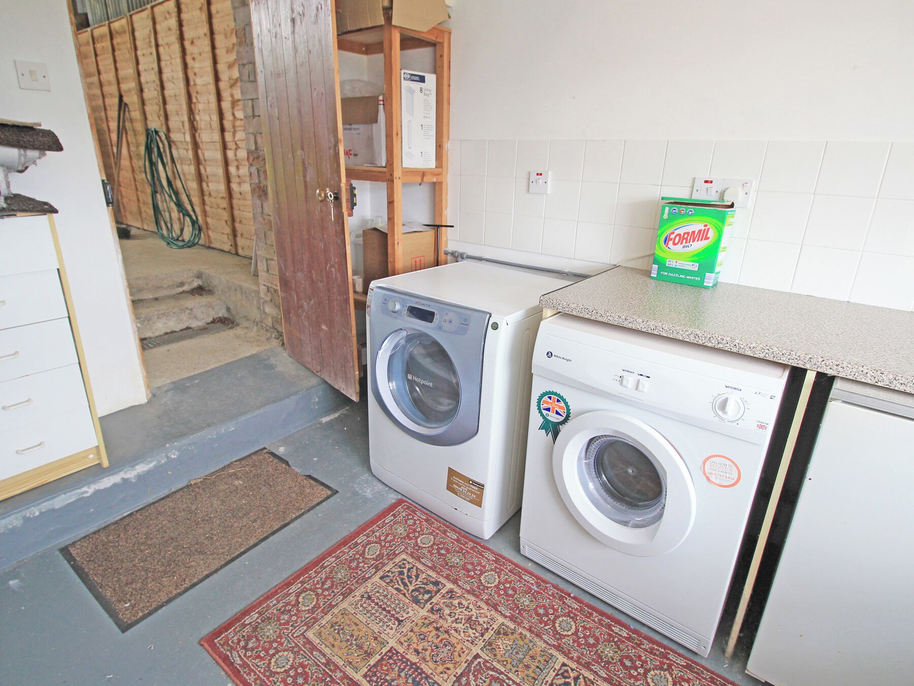 A laundry room with two washing machines, a wooden door, a doormat, and a box of Fomil soap.