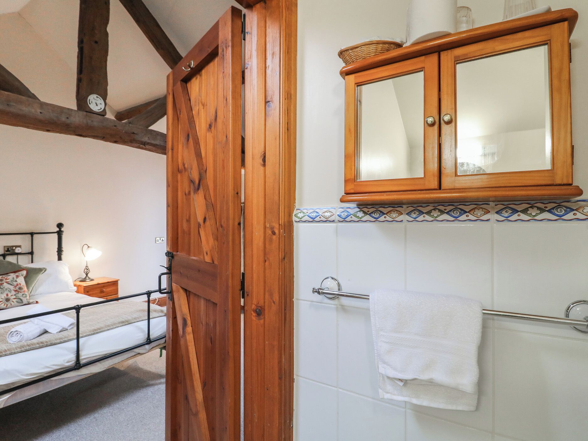 A bedroom with a wooden door, a bed with white linens, a nightstand with a lamp, and a bathroom with a towel rack and a wooden cabinet above the sink.