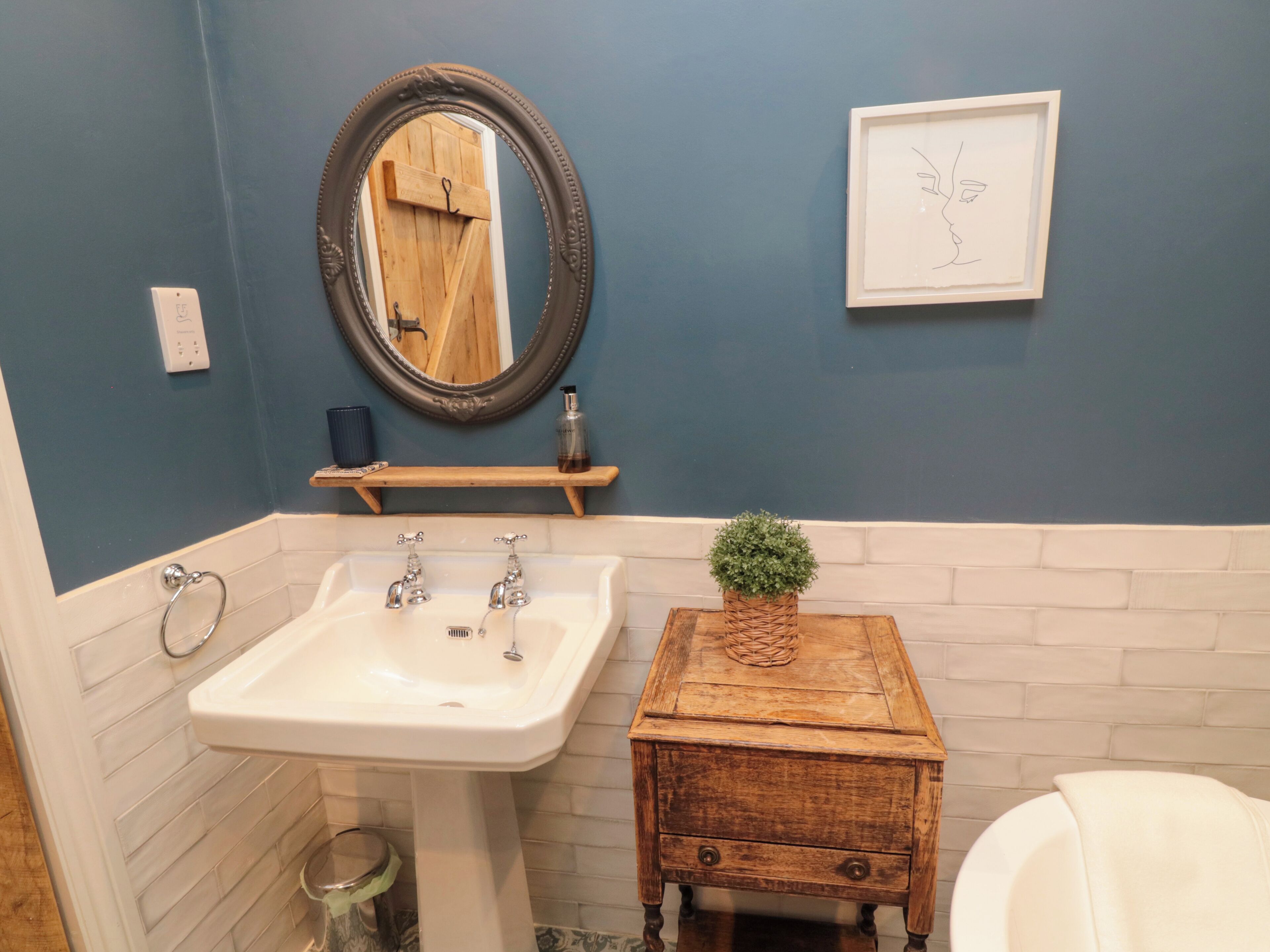 Une salle de bain avec un lavabo blanc, un meuble-lavabo en bois, un miroir, une œuvre d’art encadrée et une petite plante en pot.