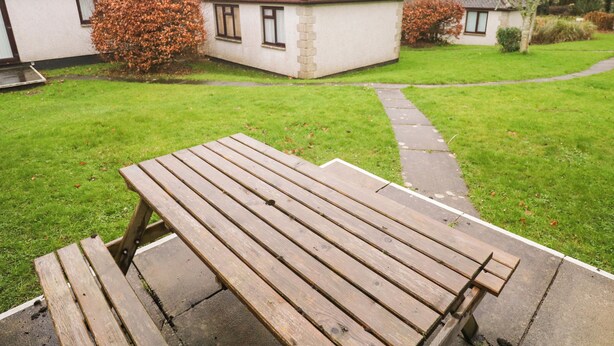 A wooden picnic table in a grassy yard with a pathway leading to a white building with a stone facade.