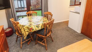 A dining area with a round table covered by a floral tablecloth, surrounded by wooden chairs. A television is mounted on the wall, and a window with a view of the outside is visible.