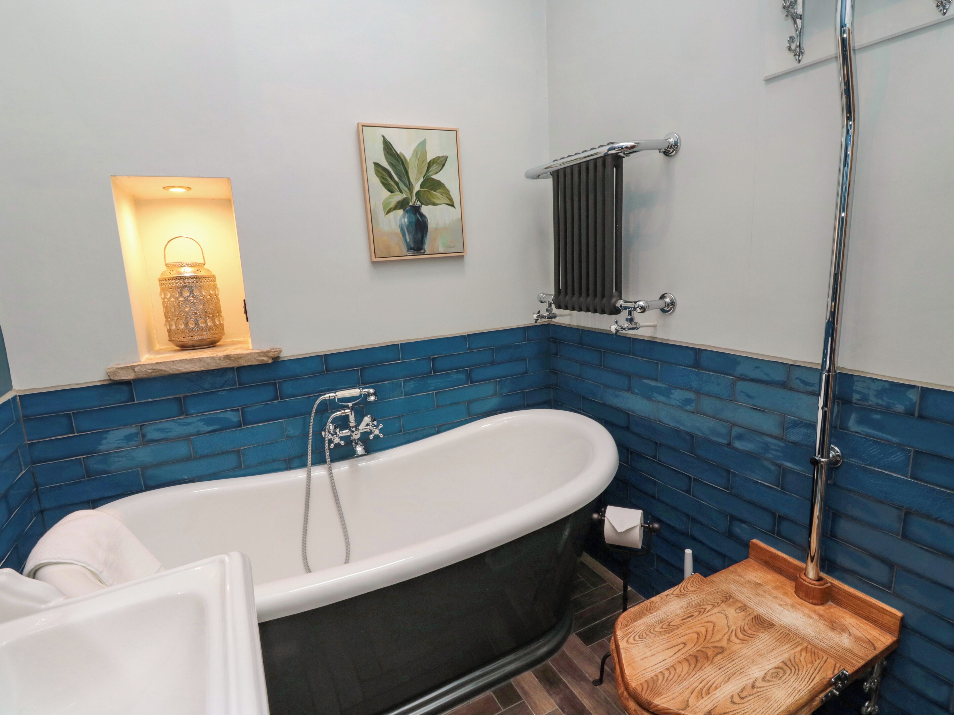 Un baño con una bañera independiente, un taburete de madera, un perchero, un radiador y una planta enmarcada en la pared.