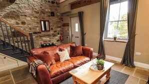 A cozy living room with a stone wall, a brown leather sofa, a wooden coffee table, and a staircase with a wooden railing. - Ash Cottage (Barnard Castle)