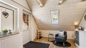 A cozy room with a sloped ceiling, a wooden floor, a black rug, a chair, a small table, a radiator, and a window with a heart decoration. - Apartment 'Krähling' with Mountain View, Shared Terrace and Wi-Fi (Schmallenberg)