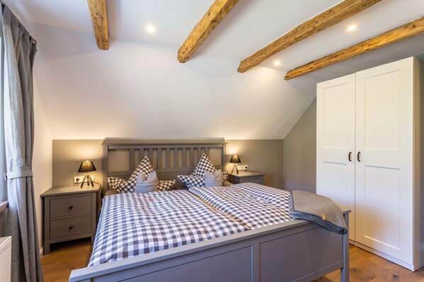 Ein Schlafzimmer mit kariertem Bettbezug, Holzbalken an der Decke, einem Nachttisch mit Lampe und einem Kleiderschrank.