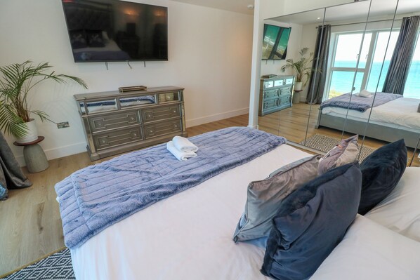 A bedroom with a bed, a dresser, a television, and a large window with a view of the sea.