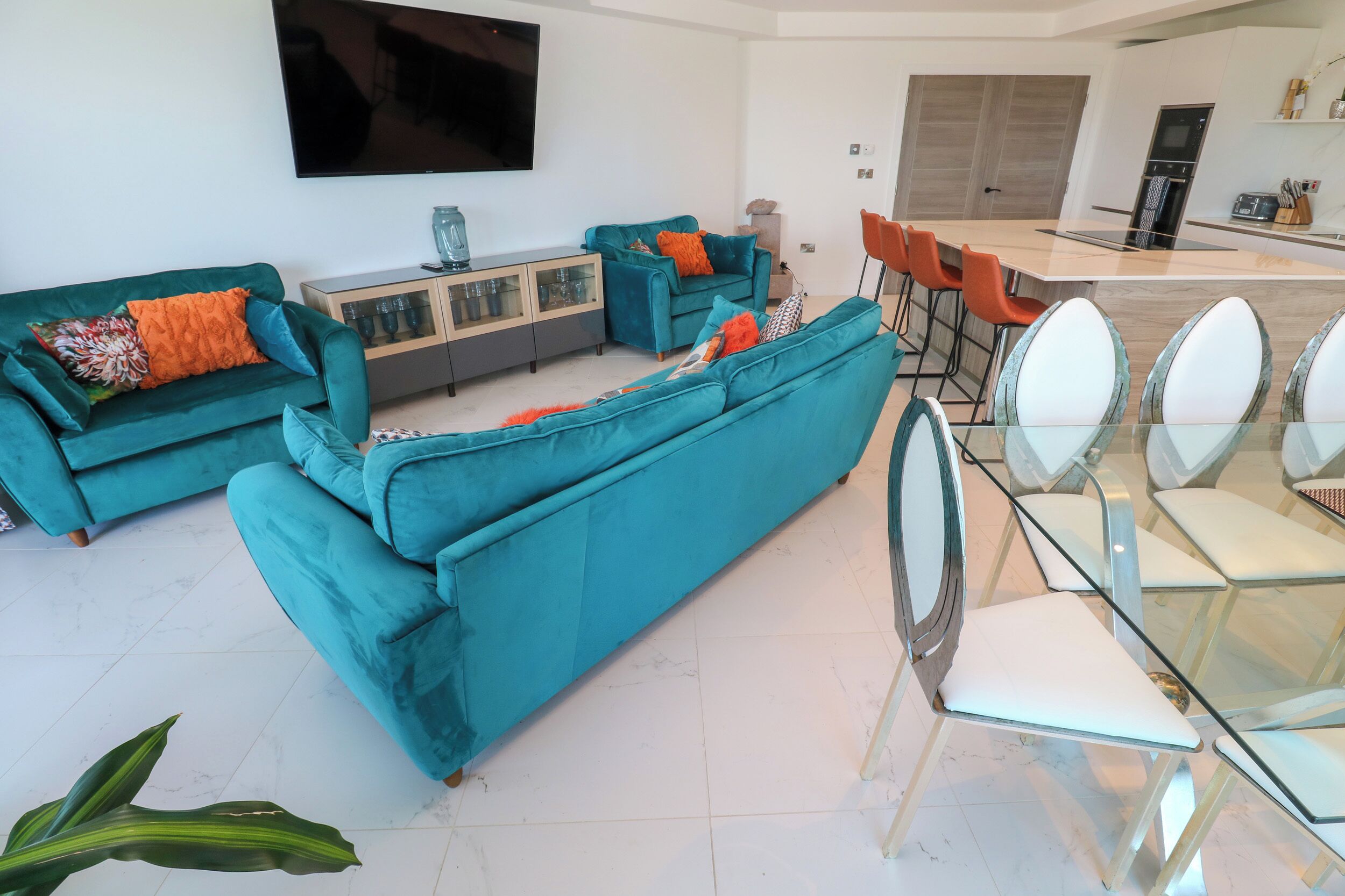 A modern living room with teal sofas, a glass dining table, and a flat-screen TV mounted on the wall.