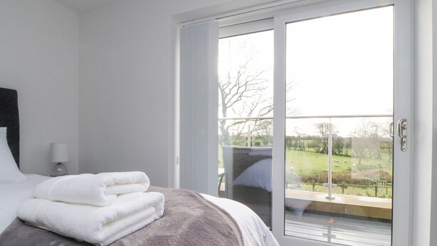 A bedroom with a bed, a nightstand, a lamp, and a view of a garden through a sliding glass door. - Apple Tree Cottage (Preston)