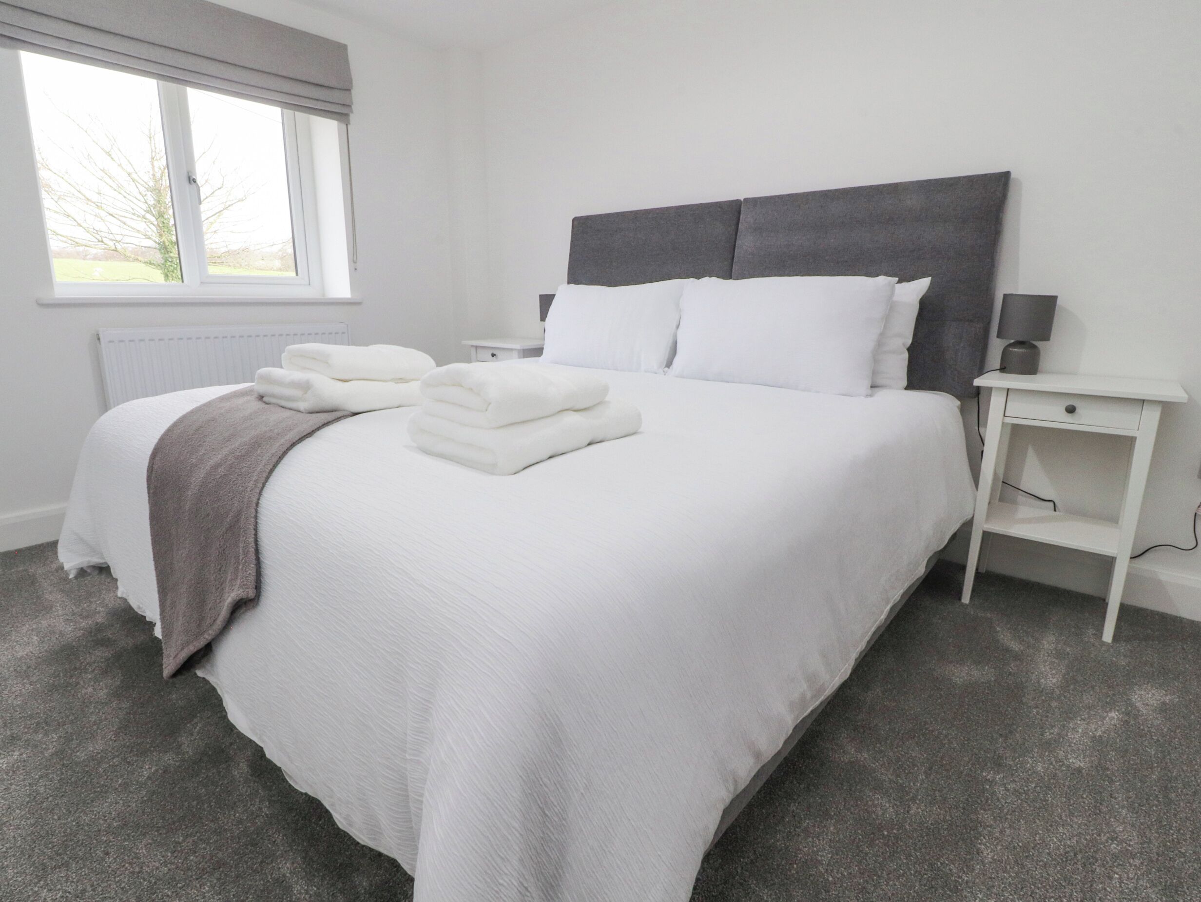 A neatly made bed with white linens and a folded towel, a bedside table with a lamp, and a window with blinds.