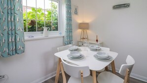 A dining area with a white table set for four, a window with seagull curtains, a white lamp, and a small plant on the windowsill. - Sea Rose (Cardigan)