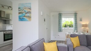 A modern living room with grey sofas and yellow cushions, a kitchen with a stove and cabinets, and a dining area with a table set for two. - Sea Rose (Cardigan)