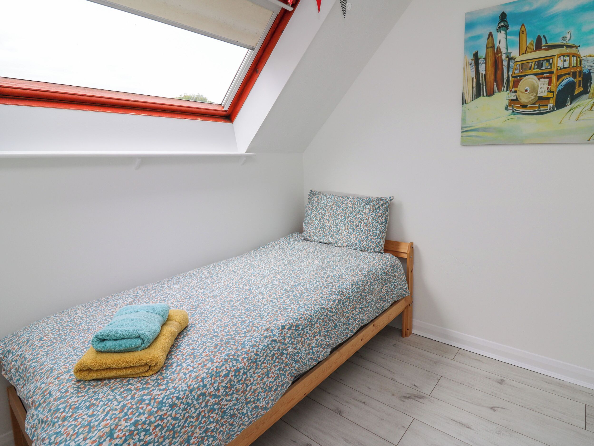 A small, single-bed room with a skylight, a painting of a coastal scene, and a wooden floor.