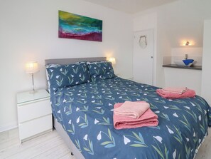 A neatly made bed with a blue floral bedspread, two bedside lamps, a painting above the bed, and a small shelf with a bowl and towel.
