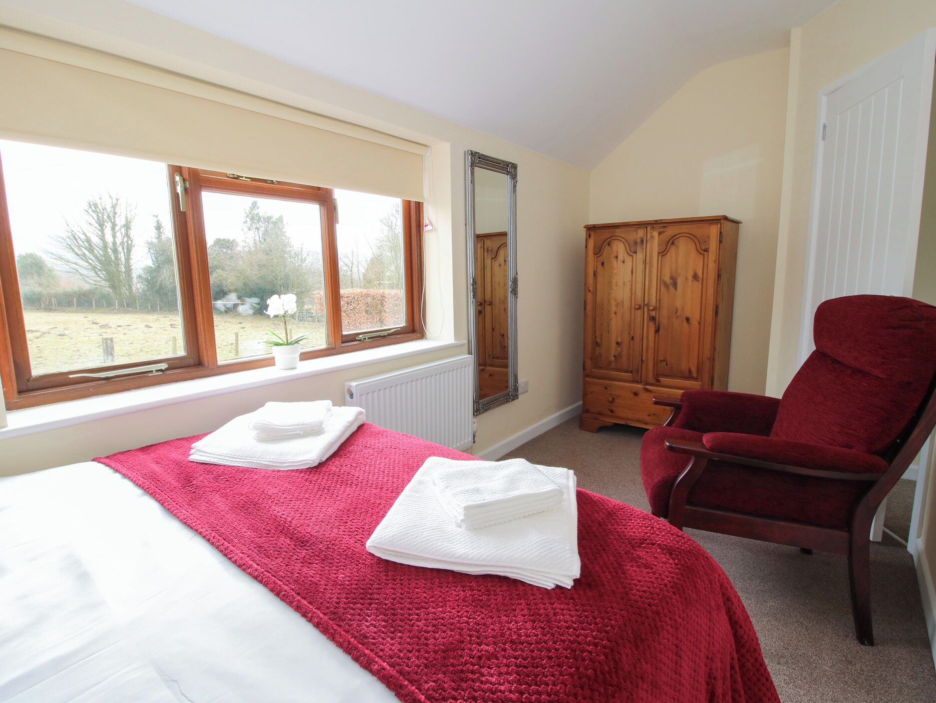 A bedroom with a bed, a red chair, a wooden wardrobe, and a window with a view of the outdoors.