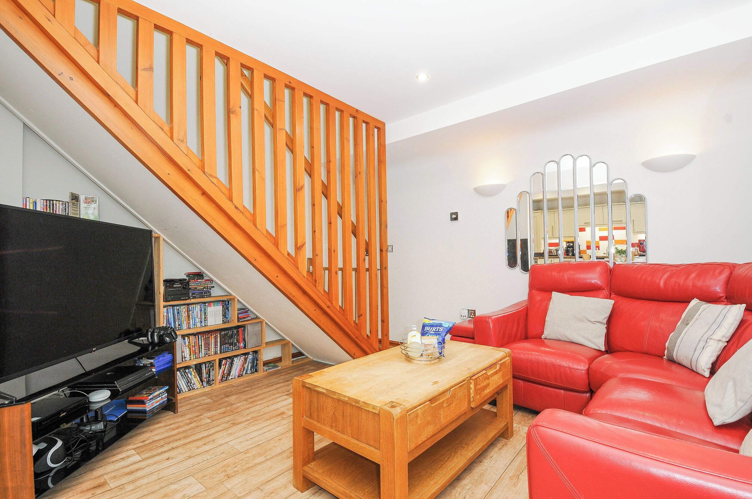 A living room with a wooden staircase, a red leather sofa, a wooden coffee table, and a bookshelf filled with books and DVDs.