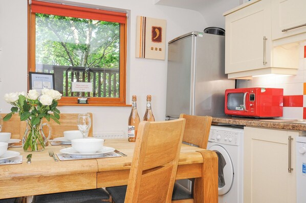 A dining area with a wooden table set for four, a window with a view of trees, a refrigerator, a microwave, and a washing machine. - Lodge Nine (Newton Abbot)