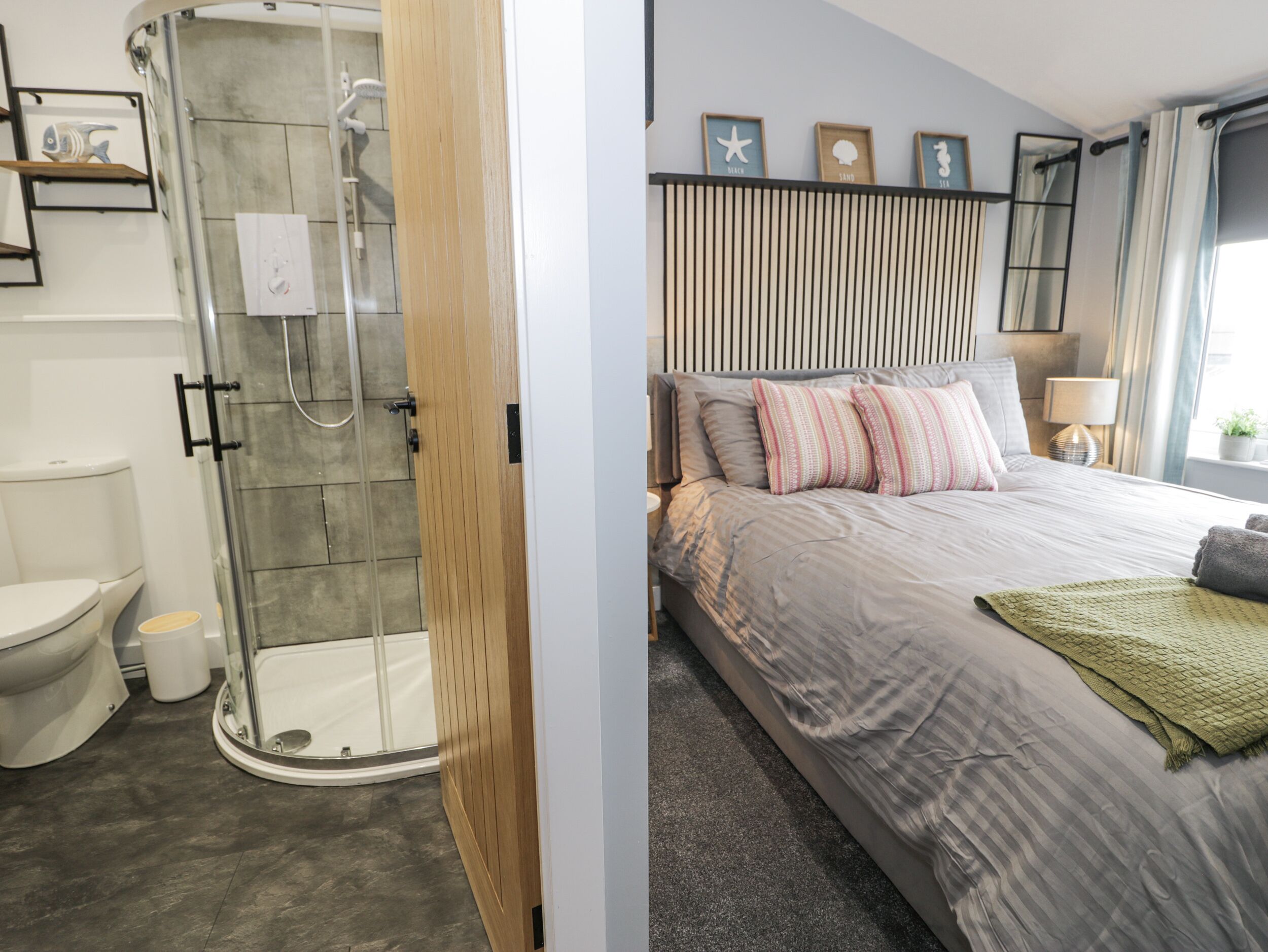 Ein modernes Badezimmer mit einer Duschkabine, einer Toilette und einem Waschbecken. Das Schlafzimmer verfügt über ein Bett mit gestreifter Bettdecke, Kissen und einer Nachttischlampe.