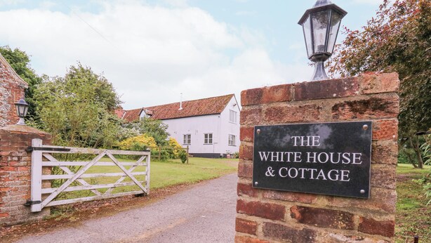 Une enseigne indiquant « The White House & Cottage », avec un mur de briques et un portail blanc menant à un bâtiment surmonté d’un toit recouvert de tuiles rouges.