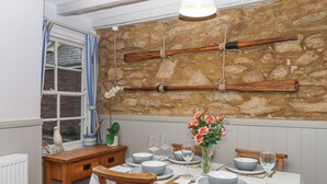 A dining area with a stone wall, wooden chairs, a table set for four, and a vase of flowers. - 3 Pebble Cottage (Brixham)