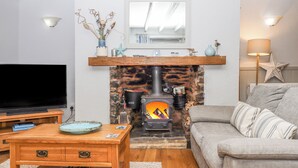 A cozy living room with a stone fireplace, a flat-screen TV, a wooden coffee table, and a grey sofa. - 3 Pebble Cottage (Brixham)