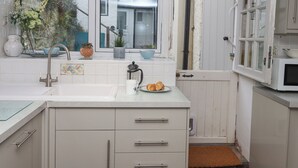 A kitchen with white cabinets, a sink, and a window overlooking a house. - 3 Pebble Cottage (Brixham)