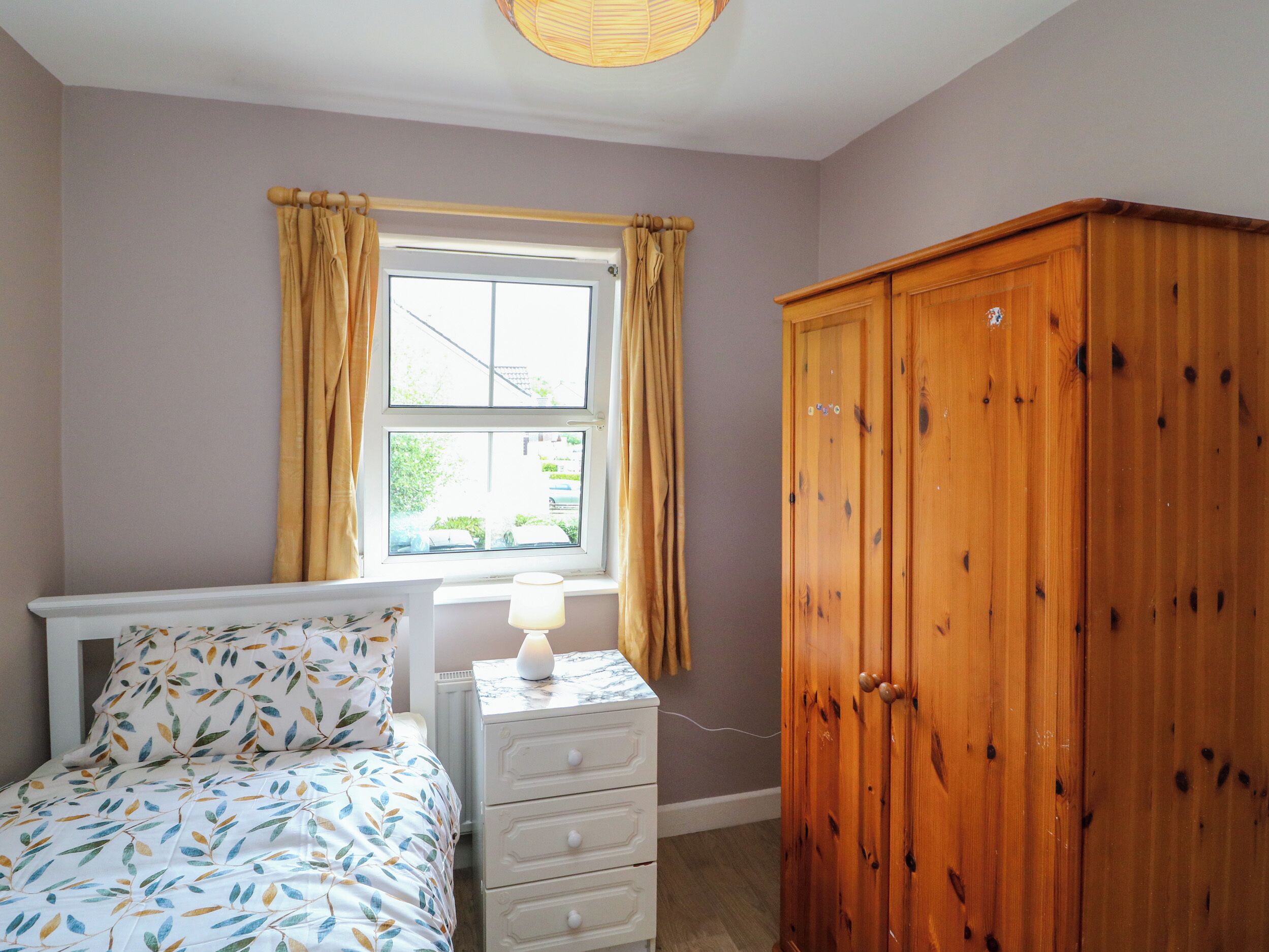 A bedroom with a bed, a nightstand, a wooden wardrobe, a window with curtains, and a lamp.
