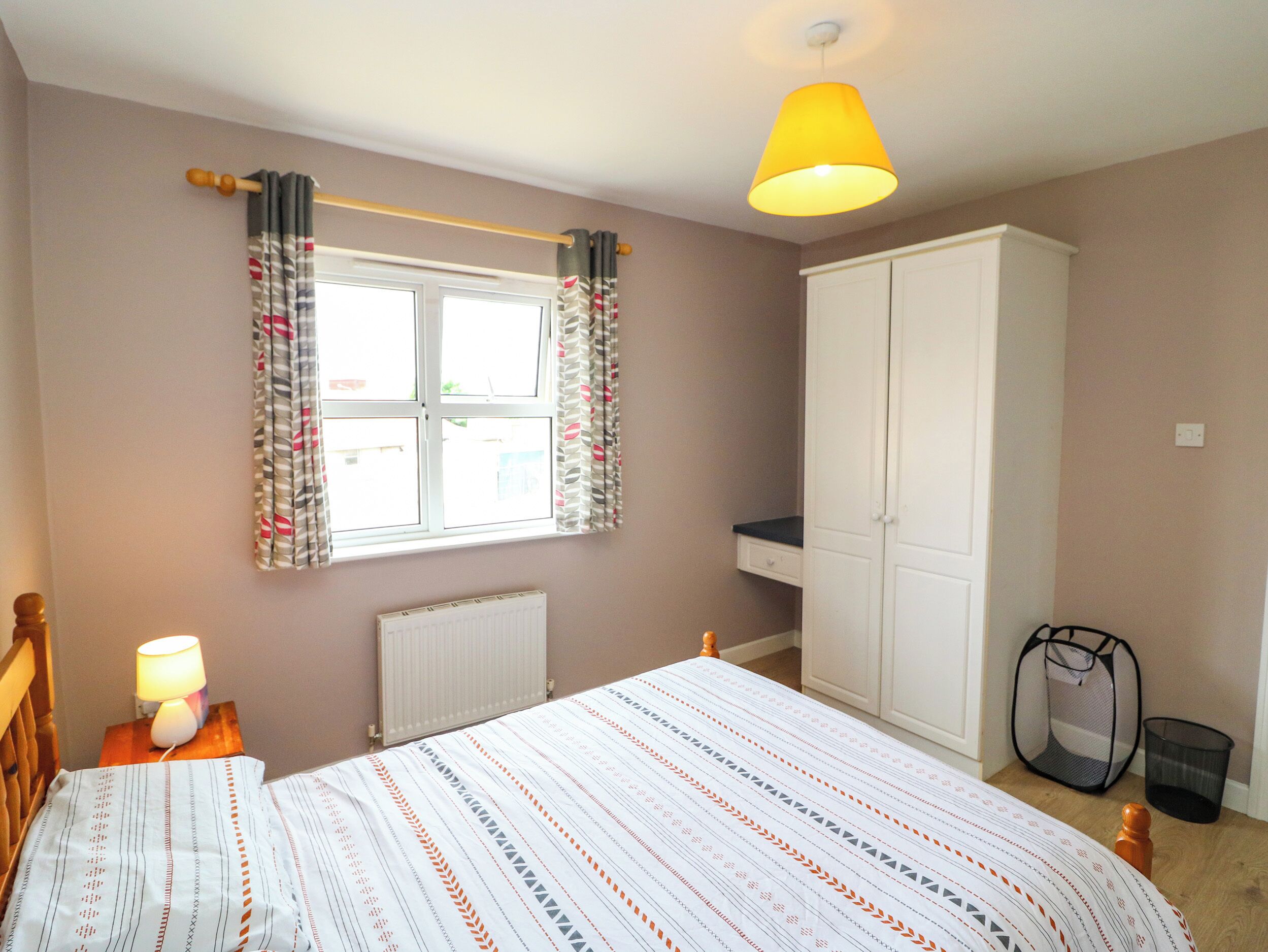 A bedroom with a bed, a window with curtains, a wardrobe, a radiator, a bedside lamp, and a hanging laundry basket.