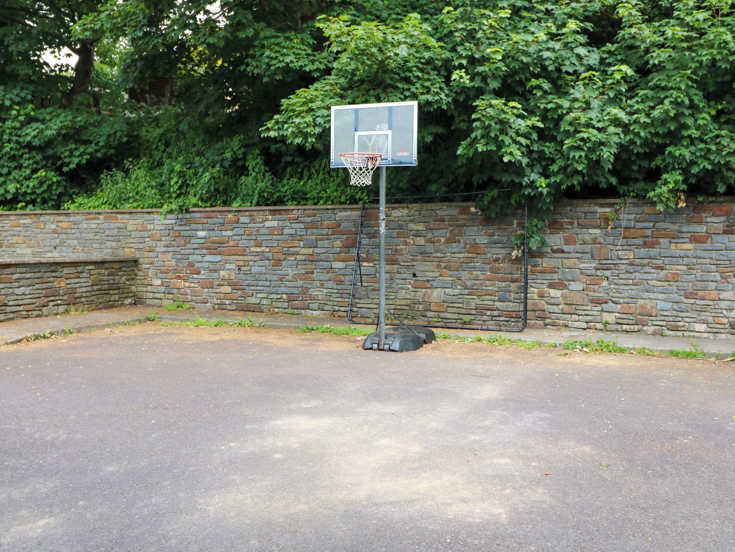 A basketball court with a metal pole and a backboard, surrounded by a stone wall and dense green foliage.