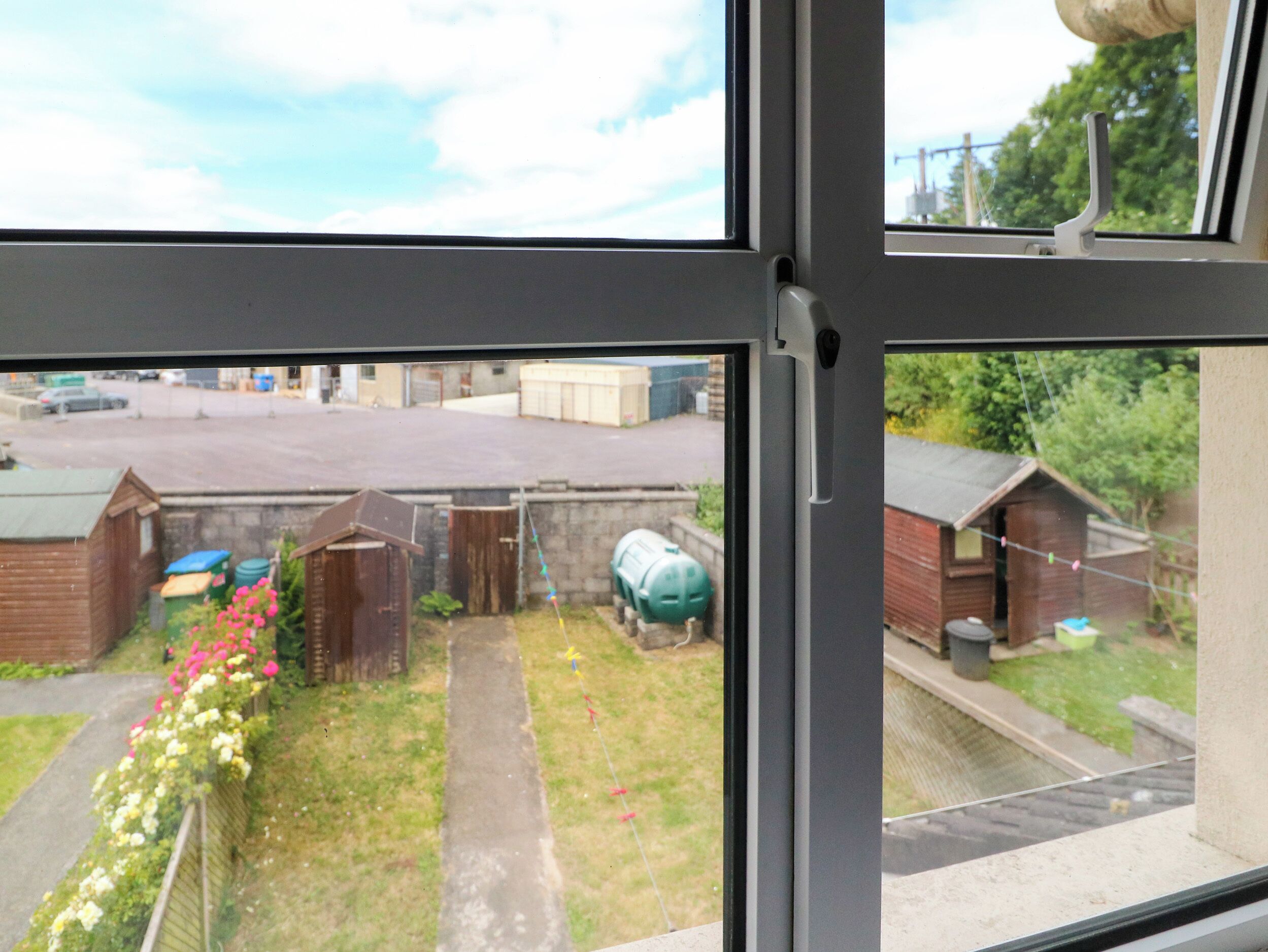 A view through a window of a residential area with a paved driveway, a small garden with flowers, and a green water tank.