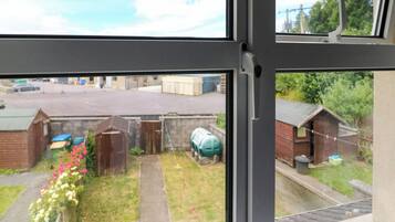 A view through a window of a residential area with a paved driveway, a small garden with flowers, and a green water tank.